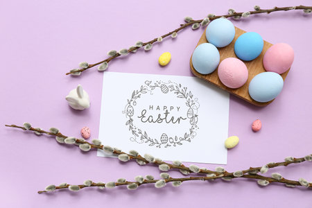 Wooden holder with Easter eggs, greeting card and willow branches on lilac backgroundの写真素材