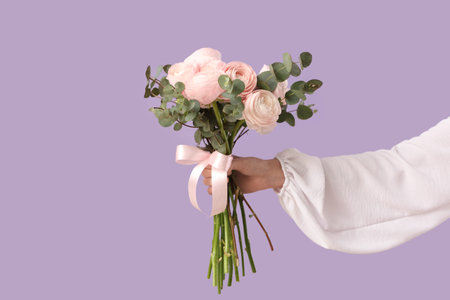 Female hand with bouquet of ranunculus flowers and eucalyptus on lilac background