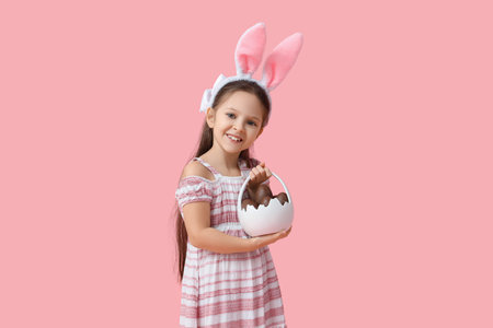 Cute little girl in bunny ears holding basket with sweet chocolate egg on pink background. Easter...