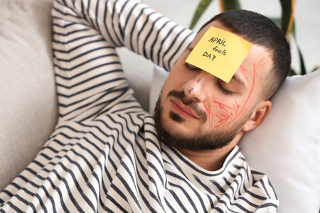 Father with paintings on his face and paper sheet sleeping on sofa at home. April fool's day prankの写真素材