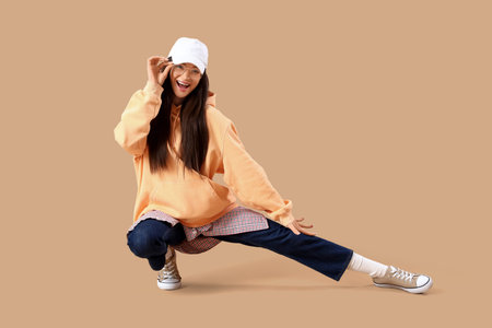 Stylish young woman in hoodie with cap on beige background