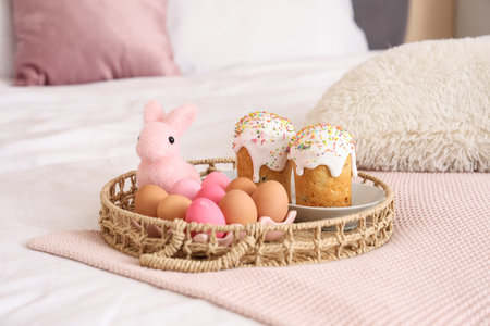 Wicker tray with Easter cakes, painted eggs and bunny on bed in festive bedroom, closeup