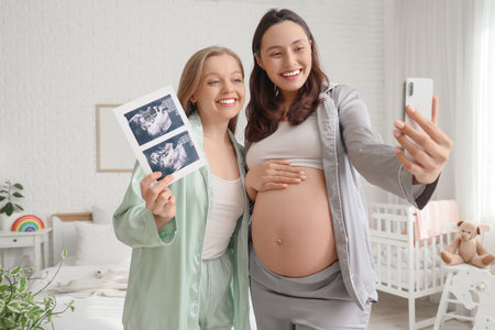 Young pregnant lesbian couple with sonogram image taking selfie in bedroomの写真素材