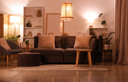 Interior of modern living room with gray sofa, armchair, pouf and glowing lamps at eveningの写真素材