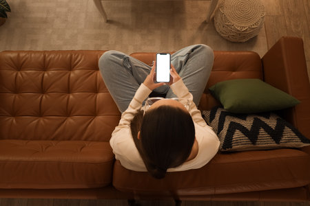 Young woman sitting on sofa and using mobile phone at home in evening. Top viewの写真素材
