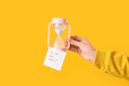 Woman holding hourglass and paper with text IF NOT NOW, WHEN? on yellow backgroundの写真素材