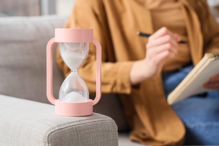 Hourglass against woman writing in notebook on sofa at home, closeup