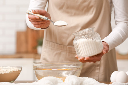 Woman adding spoon of sugar into bowl for preparing dough at table in kitchen. Closeup