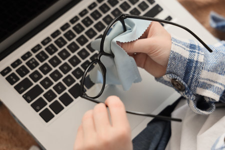 Young woman with laptop wiping new eyeglasses at home, top view