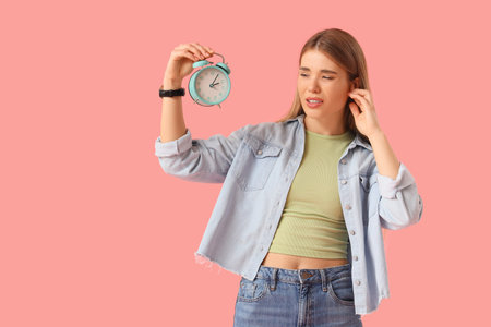 Irritated young woman with ringing alarm clock on pink background