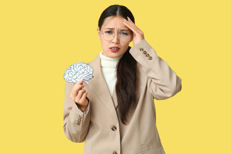 Young Asian businesswoman with paper brain suffering from headache on yellow background. Brain Injury Awareness Monthの写真素材