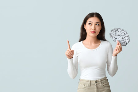 Young Asian woman with paper brain pointing up on gray background. Brain Injury Awareness Month