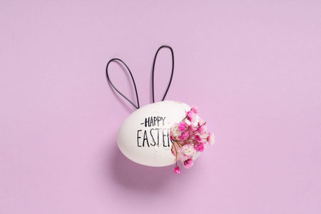 Egg shell with text HAPPY EASTER, flowers and bunny ears on lilac background