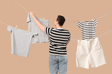 Handsome young man hanging clean clothes with wooden clothespins on beige background, back viewの写真素材