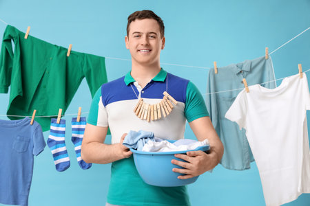 Handsome young man with hanging clean clothes, laundry basket and wooden clothespins on blue backgroundの写真素材