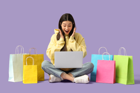 Surprised young woman with laptop and shopping bags sitting on lilac backgroundの写真素材