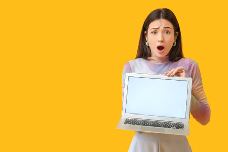 Shocked young woman with blank laptop on yellow backgroundの写真素材