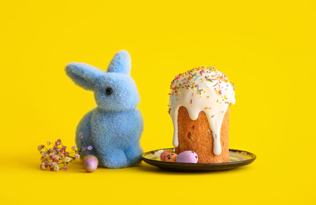 Plate with sweet Easter cake, painted eggs, toy bunny and gypsophila flowers on yellow background