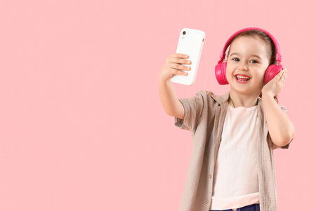 Cute little girl in headphones with mobile phone on pink backgroundの写真素材