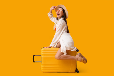 Happy young female tourist sitting on suitcase against yellow backgroundの写真素材