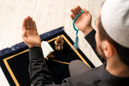 Young Muslim man with Koran and tasbih praying on prayer carpet at home, closeup. Ramadan...