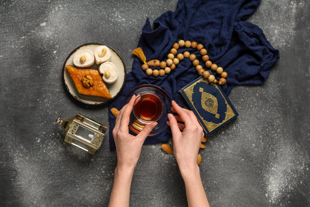 Female hands with glass of tasty Turkish tea, Koran, tasbih and sweets on gray grunge background....