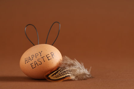 Easter egg with drawn text HAPPY EASTER, bunny ears and feather on brown backgroundの写真素材