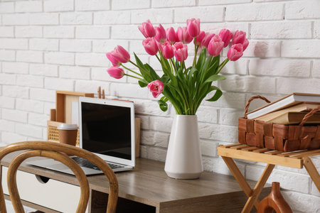 Stylish workspace with modern laptop, cup of coffee and bouquet of beautiful tulip flowers near white brick wallの写真素材