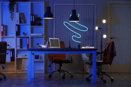 Interior of office with workplace, shelf unit and glowing lamps at nightの写真素材