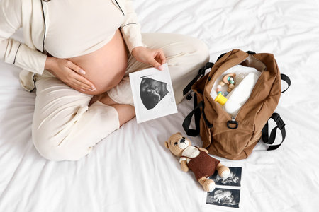 Young pregnant woman with sonogram image and maternity hospital bag on bed, top view