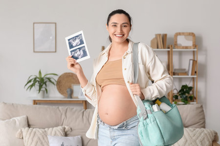 Young pregnant woman with sonogram image and maternity hospital bag at home