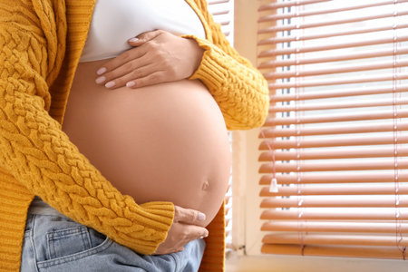 Young pregnant woman near window at home, closeup