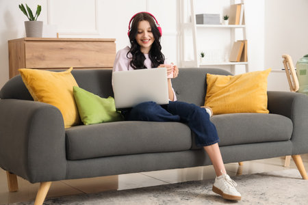 Teenage girl in headphones with cup of tea and laptop working online on sofa at homeの写真素材