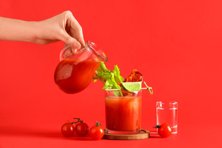 Woman pouring tomato juice from jug into glass of bloody mary cocktail with bacon and celery on red backgroundの写真素材
