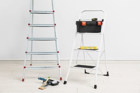 Maintenance ladders with instruments and paint tools near white wall