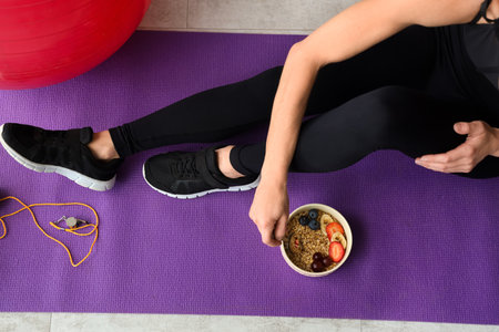 Mature sporty woman with bowl of nutritious oatmeal and fruits sitting on yoga mat, top viewの写真素材