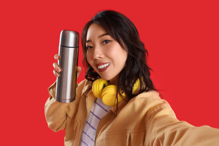 Young Asian woman with thermos taking selfie on red background, closeup