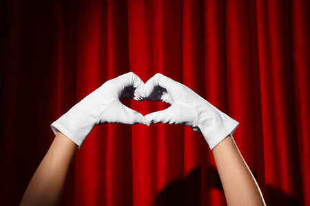 Female hands in gloves making heart gesture against red curtainの写真素材