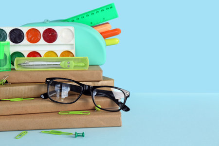 Stack of books with different stationery supplies and eyeglasses on blue background