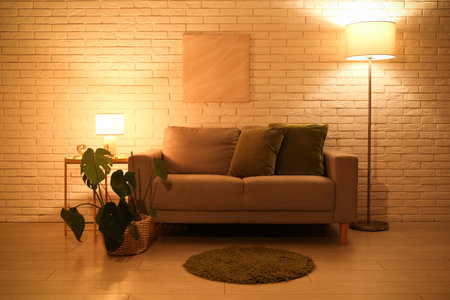 Interior of night living room with sofa, glowing lamps and houseplant on stool near white brick wallの写真素材