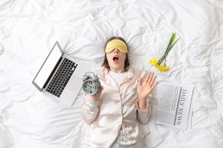 Shocked young woman in pajamas with alarm clock and laptop lying on bed, top view