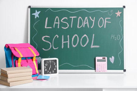 Table with backpack, books, clock and text LAST DAY OF SCHOOL on blackboard near light wallの写真素材