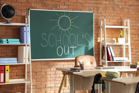 Blackboard with text SCHOOL'S OUT and drawn sun on brick wall in classroom interiorの写真素材