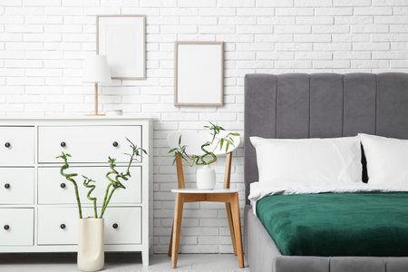 Interior of stylish bedroom with cozy bed, dresser and bamboo stems on chair