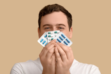 Happy young man covering mouth with blisters of pills on beige background, closeupの写真素材