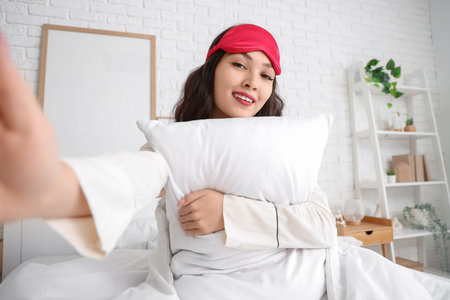 Beautiful young woman with sleep mask and pillow taking selfie in bed at home