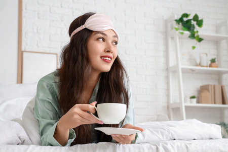 Beautiful young woman with sleep mask and cup of coffee lying in bed at homeの写真素材