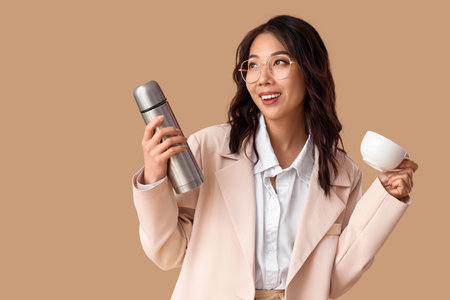 Young Asian businesswoman with thermos and cup on beige backgroundの写真素材