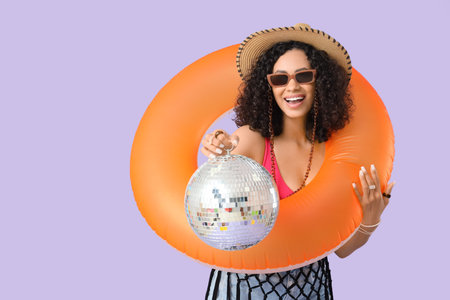 Stylish woman with inflatable ring and disco ball on lilac background