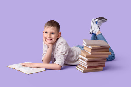 Cute little boy with stack of books lying on lilac backgroundの写真素材